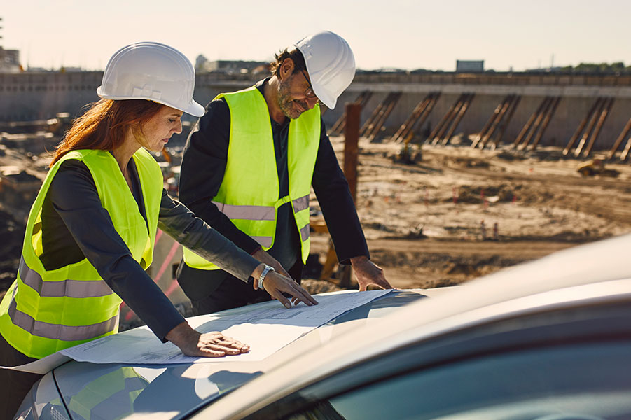 Zwei Personen in Sicherheitshelmen und Warnwesten betrachten Baupläne auf einer Baustelle.