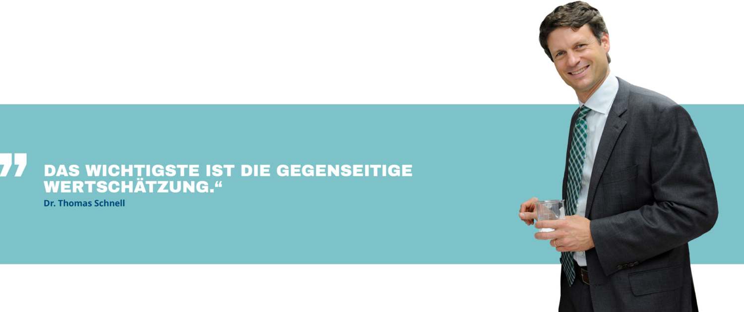 T. Schnell in einem Anzug hält ein Glas und steht vor einem Zitat: "Das Wichtigste ist die gegenseitige Wertschätzung." Links neben ihm ist ein blauer Hintergrund.