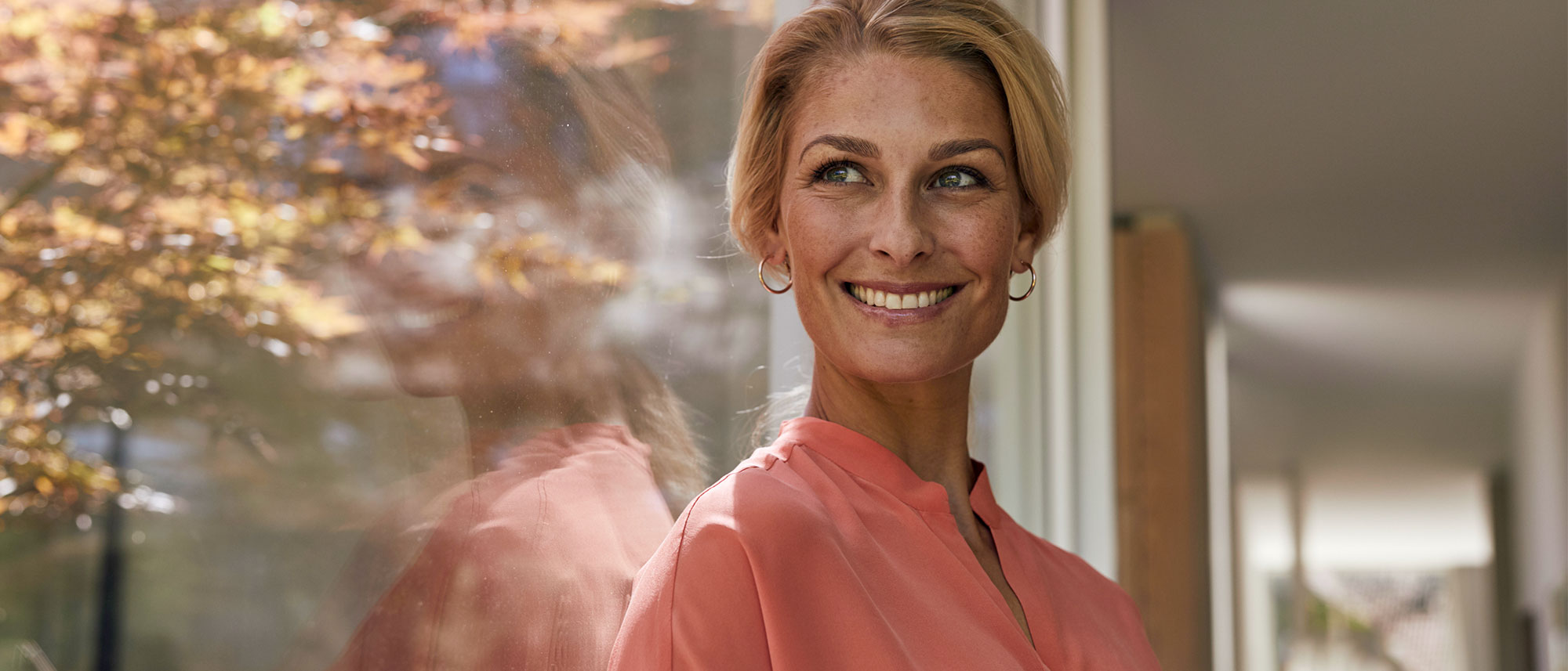Eine lächelnde Frau in einem rosa Oberteil steht neben einem Fenster, durch das herbstliche Bäume zu sehen sind.