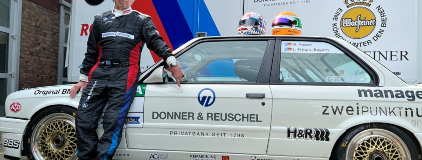 Ein Mann in Rennanzug steht neben einem weißen Rennwagen mit der Aufschrift "DONNER & REUSCHEL". Im Hintergrund ist ein BMW-Transporter zu sehen.