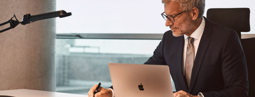 Ein Mann in einem Anzug sitzt an einem Schreibtisch und arbeitet an einem Laptop. Er hält einen Stift in der Hand und betrachtet Dokumente. Im Hintergrund ist ein Fenster zu sehen.