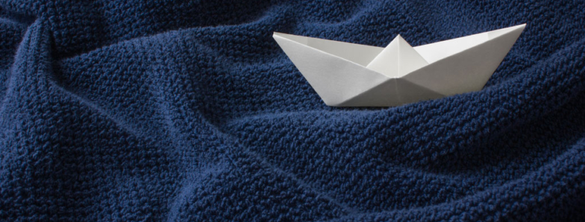 Ein weißes Papierboot liegt auf einem dunkelblauen, strukturierten Stoff.