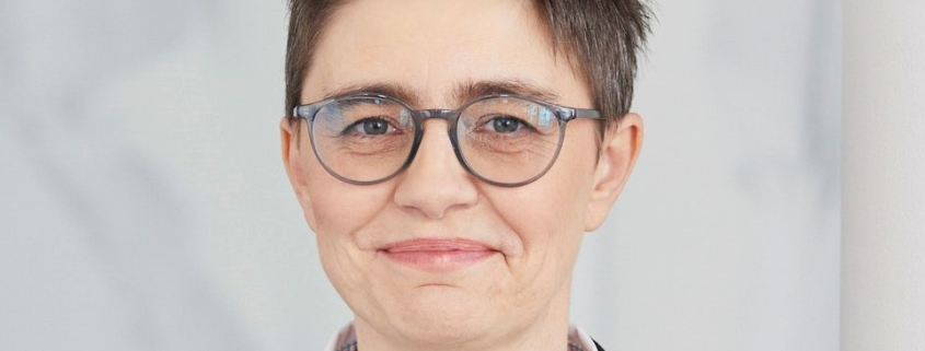 Eine Person mit kurzen Haaren und Brille trägt einen schwarzen Anzug und ein weißes Hemd. Sie lächelt leicht und steht vor einem hellen Hintergrund.