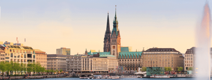 Blick auf das Hamburger Rathaus und die Binnenalster bei Sonnenuntergang, mit einem Springbrunnen im Vordergrund.
