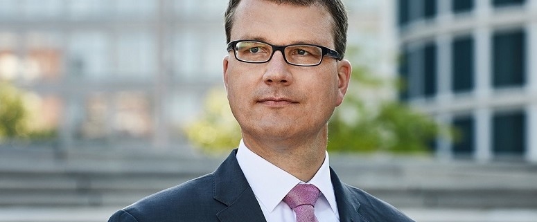 Carsten Mumm in Anzug und Krawatte steht draußen vor einem modernen Gebäude. Er trägt eine Brille und schaut zur Seite.
