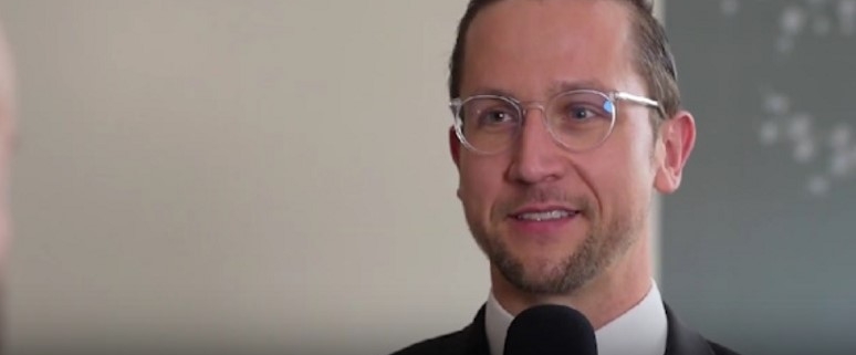 Tobias Regele mit Brille und Anzug spricht in ein Mikrofon.