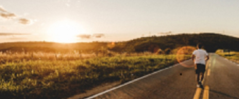 Ein Mensch joggt auf einer Landstraße bei Sonnenuntergang, umgeben von grünen Feldern und Hügeln.