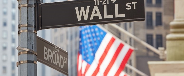 Straßenschilder von Wall Street und Broad Street mit unscharfer US-Flagge im Hintergrund.