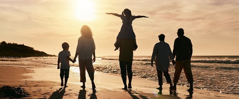 Eine Familie spaziert am Strand bei Sonnenuntergang. Ein Kind sitzt auf den Schultern eines Erwachsenen, während die anderen Hand in Hand gehen.