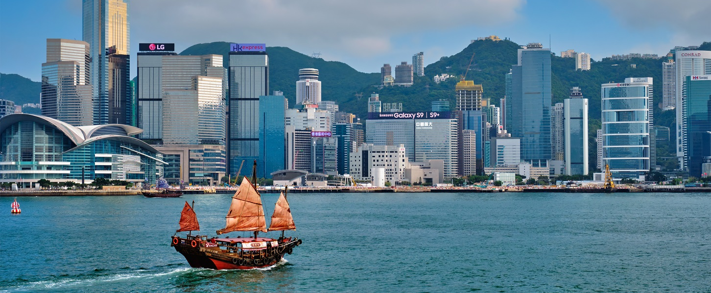 Skyline von Hongkong mit traditionellen Segelboot im Vordergrund und modernen Wolkenkratzern im Hintergrund.