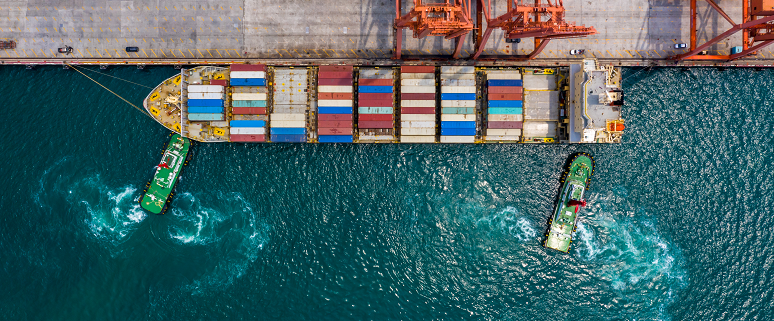 Luftaufnahme eines Frachtschiffs im Hafen, beladen mit bunten Containern. Zwei Schlepperboote manövrieren das Schiff.