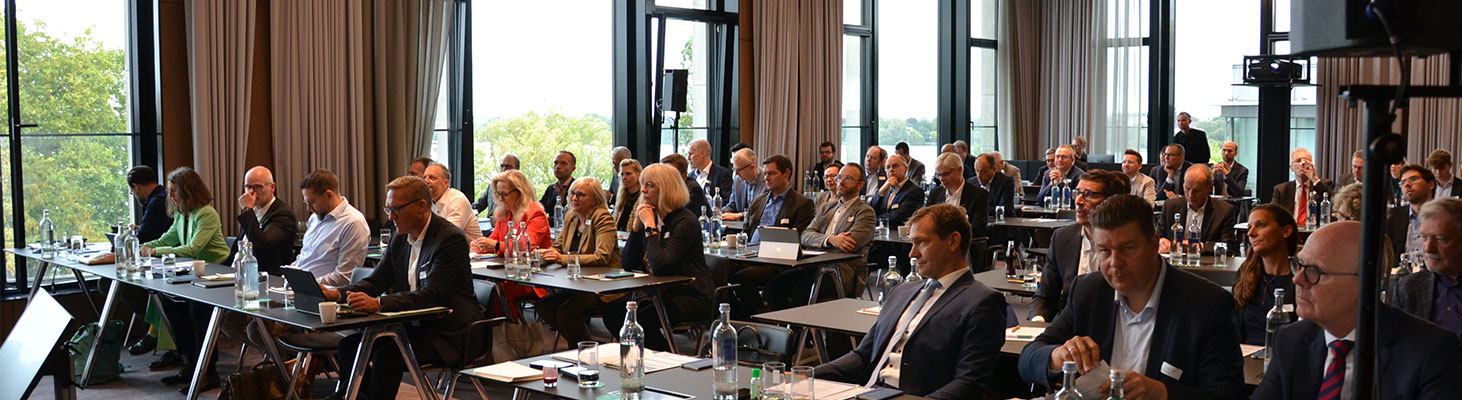Eine Gruppe von etwa 40 Erwachsenen in Businesskleidung sitzt an Tischen mit Laptops und Wasserflaschen und verfolgt aufmerksam eine Präsentation in einem Konferenzraum. Große Fenster bieten einen Blick ins Grüne und schaffen eine angenehme Atmosphäre.