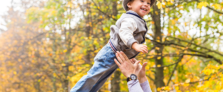 Ein lachendes Kleinkind wird in einem Herbstwalt von erwachsenen Armen leicht in die Luft geworfen und wieder aufgefangen.