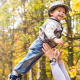 Ein lachendes Kleinkind wird in einem Herbstwalt von erwachsenen Armen leicht in die Luft geworfen und wieder aufgefangen.