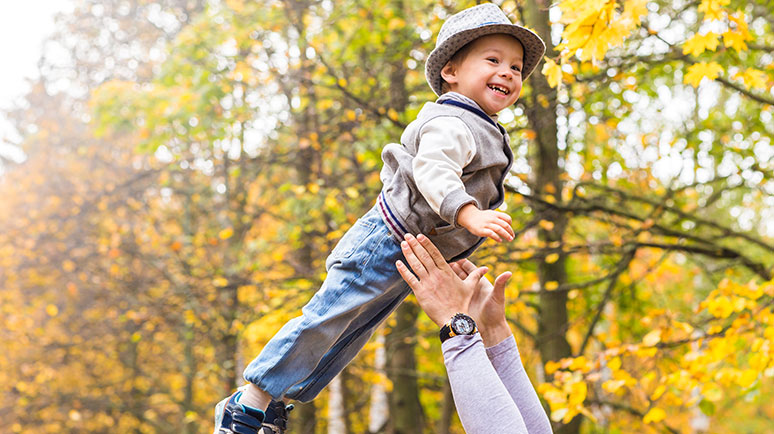 Ein lachendes Kleinkind wird in einem Herbstwalt von erwachsenen Armen leicht in die Luft geworfen und wieder aufgefangen.