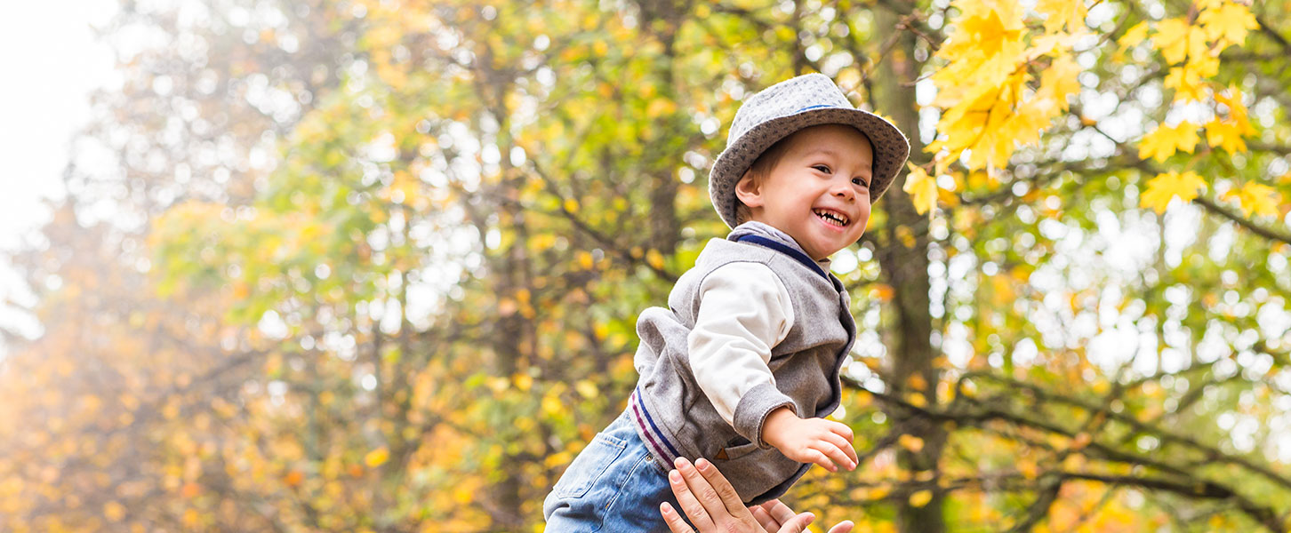 Ein lachendes Kleinkind wird in einem Herbstwalt von erwachsenen Armen leicht in die Luft geworfen und wieder aufgefangen.