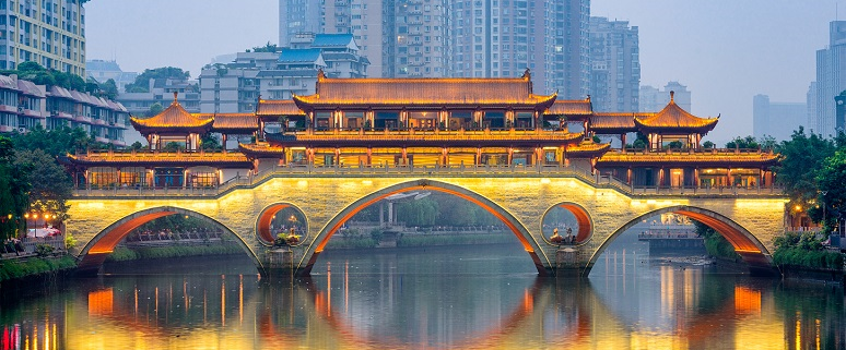 Chinesische Brücke in Chengdu bei Dämmerung, beleuchtet mit goldenen Lichtern, spiegelt sich im Wasser, umgeben von modernen Gebäuden.