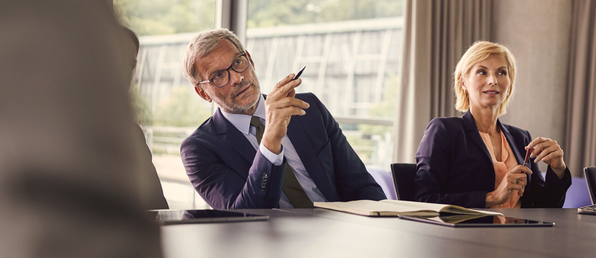 Ein Mann und eine Frau in Business-Kleidung sitzen in einem modernen Konferenzraum. Der Mann hält einen Stift und scheint aufmerksam zuzuhören. Im Hintergrund sind große Fenster mit Blick auf eine grüne Landschaft.