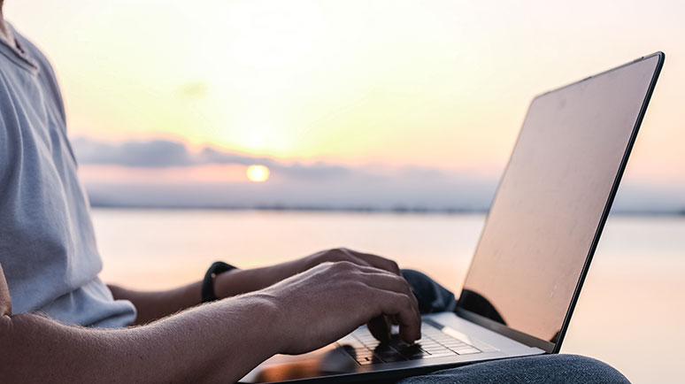 Auf dem Bild sieht man wie eine Person vor einem See an einem Laptop arbeitet