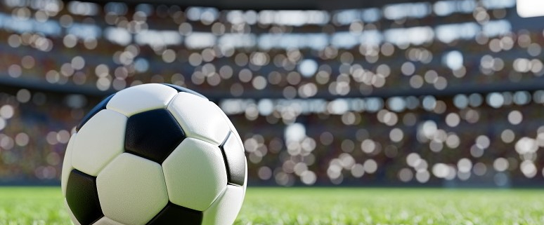Ein Fußball liegt auf einem grünen Spielfeld in einem Stadion. Im Hintergrund sind unscharfe Zuschauerränge zu sehen.