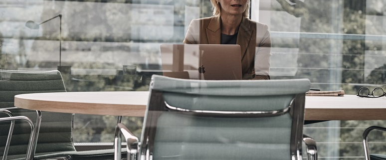 Eine Frau sitzt an einem runden Konferenztisch mit einem Laptop vor sich. Im Hintergrund sind Glaswände und moderne Büromöbel zu sehen.