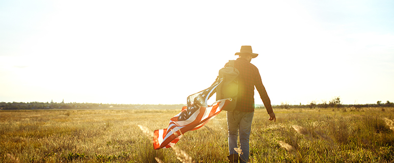 Eine Person mit Hut geht durch ein Feld und trägt eine amerikanische Flagge über der Schulter.