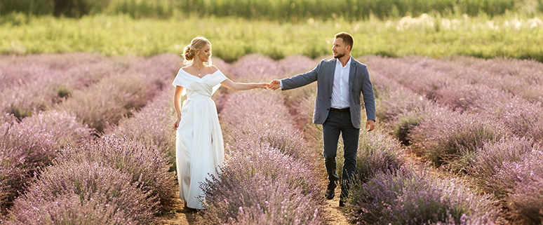 Ein Brautpaar spaziert Hand in Hand durch ein Lavendelfeld. Die Braut trägt ein weißes Kleid, der Bräutigam einen blauen Anzug. Im Hintergrund sind grüne Bäume und ein klarer Himmel zu sehen.
