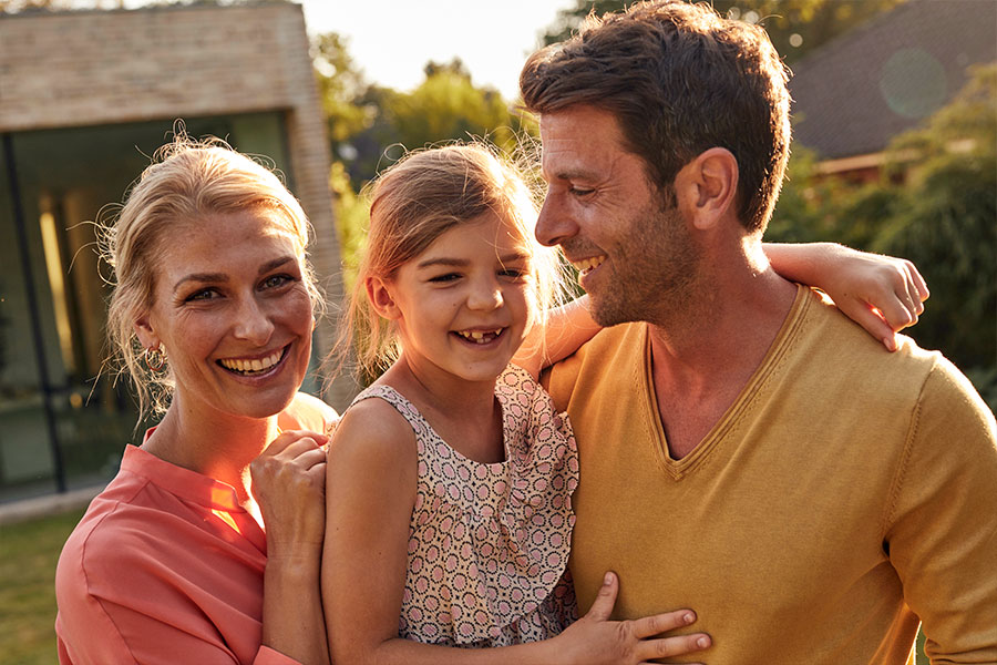 Eine Frau und ein Mann sind mit ihrer Tochter im Garten und lachen. Im Hintergrund ist ihr Haus zu sehen