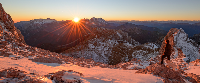 Eine Person steht auf einem schneebedeckten Berg und blickt in die Ferne, während die Sonne über den Bergen am Horizont aufgeht.